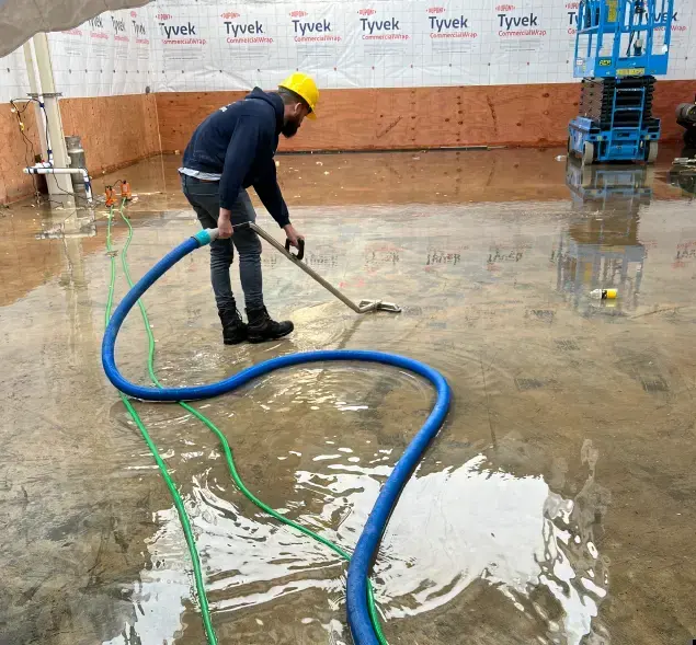 Worker extracting standing water from a flooded floor with a long blue hose in Kernersville